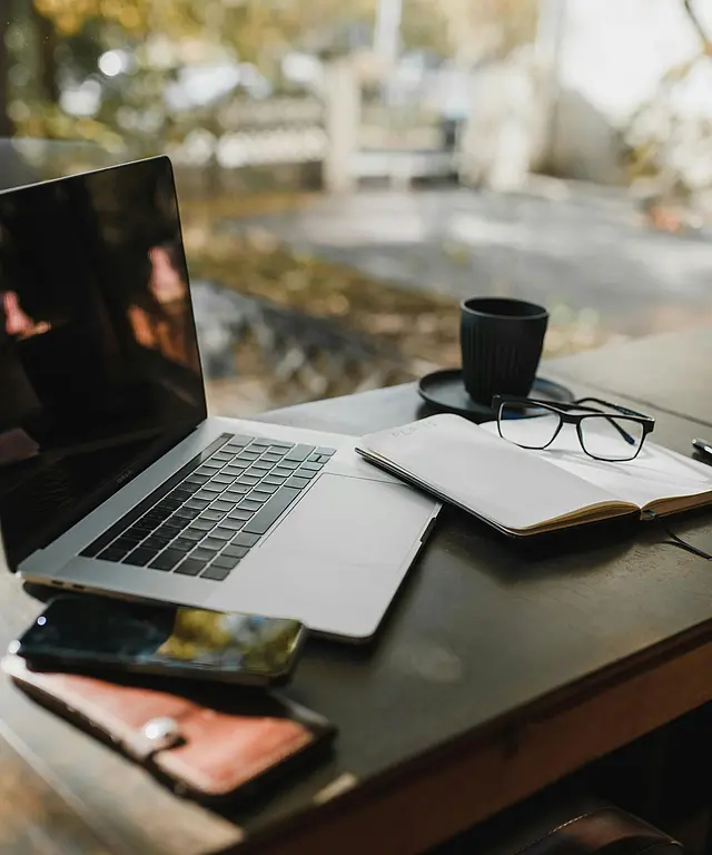 Laptop‑Arbeitsplatz mit Notizbuch, Brille und Kaffee an einem Fensterplatz. Heller, moderner Arbeitsplatz für Fokus und kreatives Arbeiten.