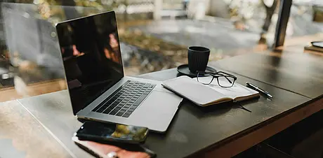 Laptop‑Arbeitsplatz mit Notizbuch, Brille und Kaffee an einem Fensterplatz. Heller, moderner Arbeitsplatz für Fokus und kreatives Arbeiten.