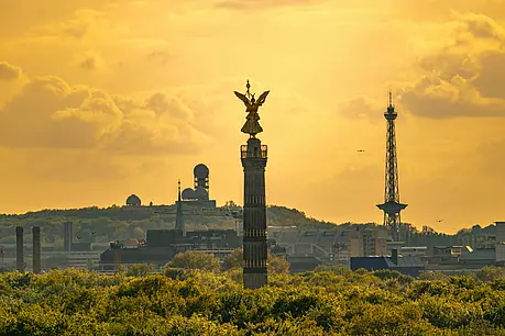 Panorama bei goldenem Himmel mit der Siegessäule im Vordergrund und weiteren Berliner Wahrzeichen wie modernem Funkturm und Radaranlagen im Hintergrund.