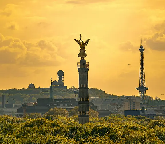 Panorama bei goldenem Himmel mit der Siegessäule im Vordergrund und weiteren Berliner Wahrzeichen wie modernem Funkturm und Radaranlagen im Hintergrund.