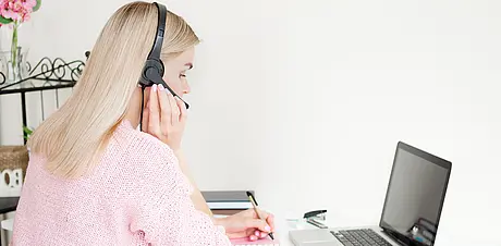 Eine Person sitzt am Schreibtisch mit Laptop und Kopfhörern und macht sich Notizen in ein Notizbuch.