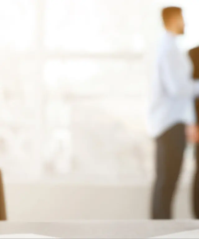 Unscharfe Gruppe von Geschäftsleuten im Gespräch vor großen Fenstern, im Vordergrund ein Schreibtisch mit Laptop – modernes Büro‑ und Teamwork‑Motiv.