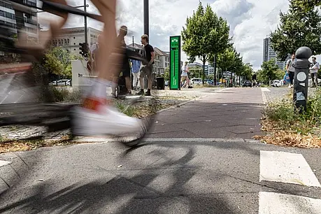 Schnell vorbeifahrendes Fahrrad im Vordergrund eines städtischen Radwegs; im Hintergrund stehen Personen, Bäume, Verkehr und moderne Gebäude.