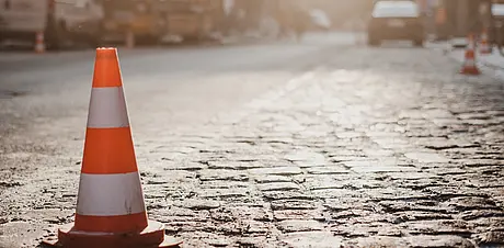 Ein orange‑weißer Verkehrskegel steht auf einer gepflasterten Straße im warmen Sonnenlicht, im Hintergrund unscharfe Baustellenszene.